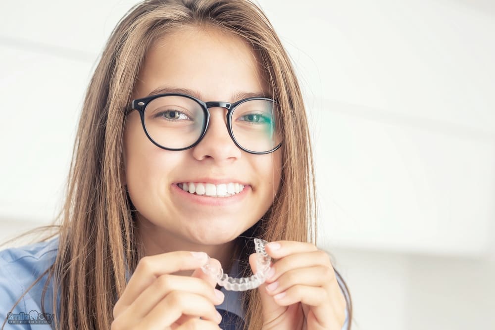 A young woman with glasses holding a clear aligner and smiling, showing an alternative method for how to fix overbite in Sunnyvale, TX.