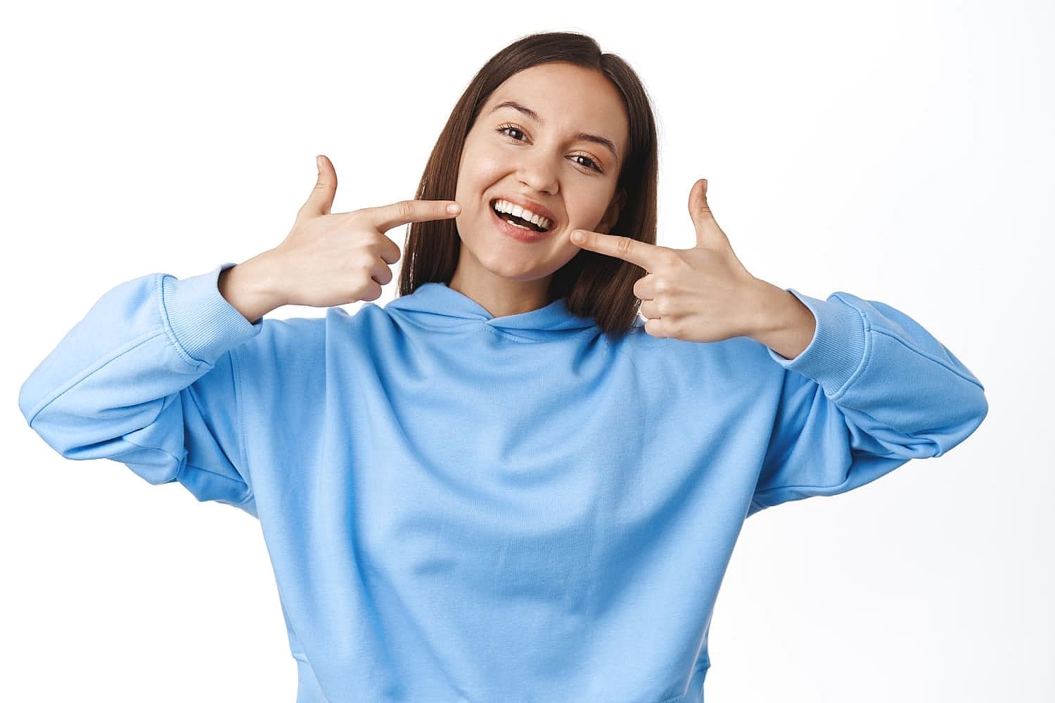 A woman in a blue hoodie smiles and points at her metal braces with both hands, proudly showcasing her orthodontic treatment at Smile City Orthodontics in Sunnyvale TX, against a white background.