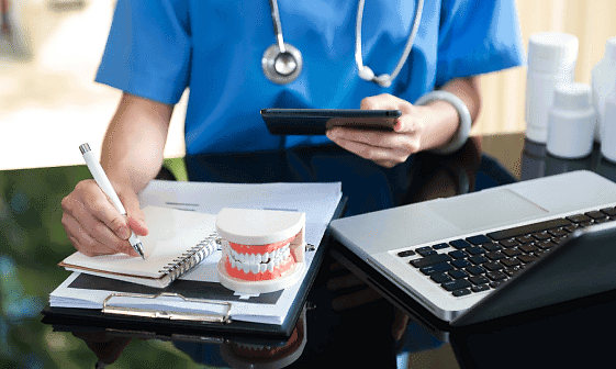 At Smile City Orthodontics in Sunnyvale TX, a dentist in scrubs reviews orthodontic insurance plans at a desk with a dental model, clipboard, laptop, and medicine bottles.