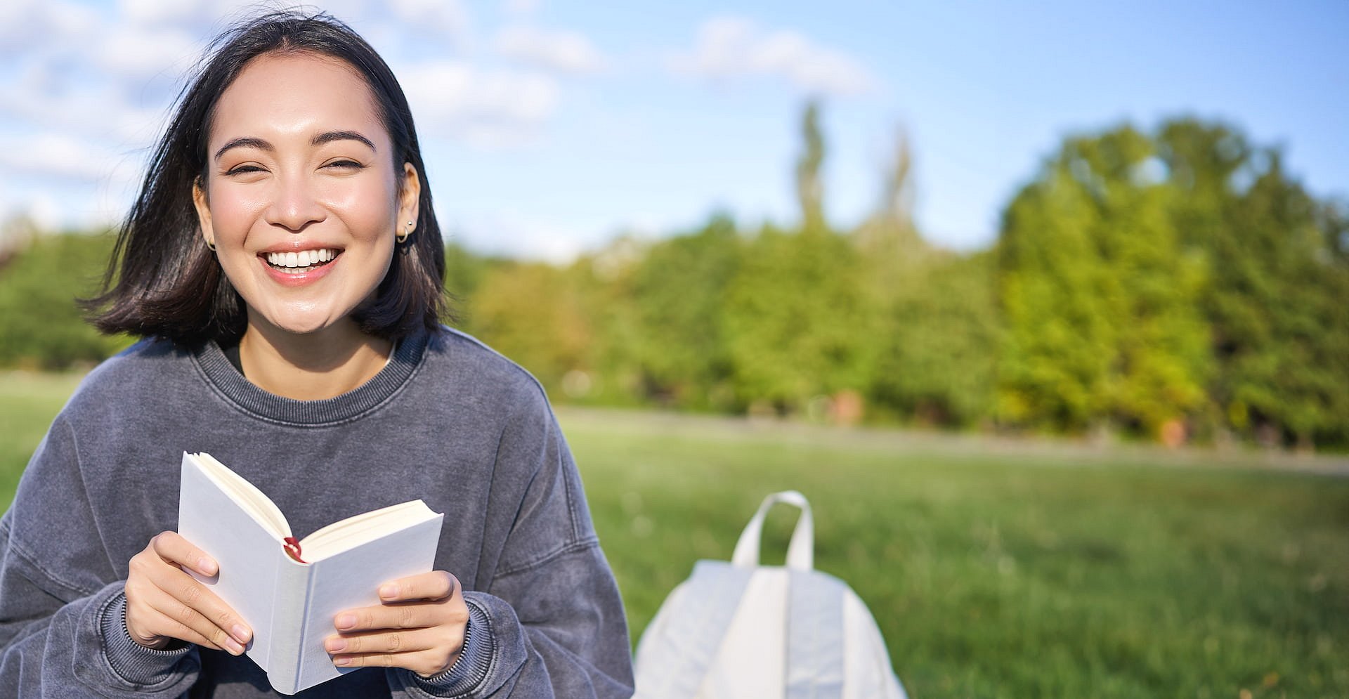 A young woman with metal braces, treated at Smile City Orthodontics in Sunnyvale TX, smiles as she sits on the grass in a park with an open book, her backpack by her side and trees in the background.