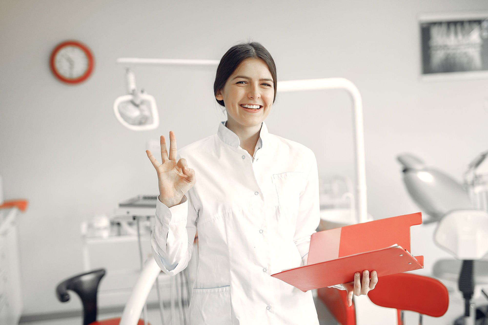 In Sunnyvale, TX, a woman in a white medical coat smiles and gives an "OK" gesture at Smile City Orthodontics, highlighting the rewarding experience of working there.