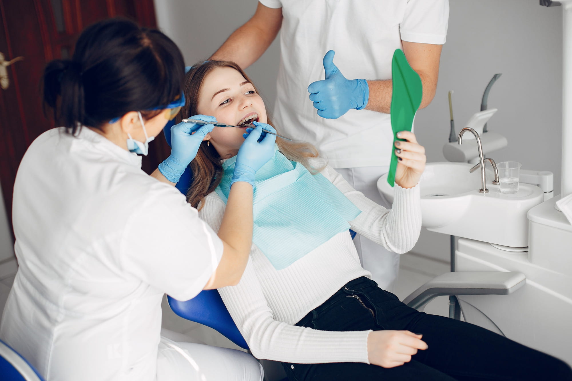 At Smile City Orthodontics in Sunnyvale TX, a dental professional adjusts a patient's braces and Herbst Appliance as the patient holds a green mirror, with another staff member nearby.
