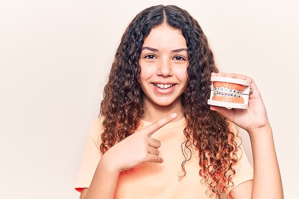 Smile City Orthodontics At Smile City Orthodontics in Sunnyvale TX, a young person with long curly hair smiles and points to a dental model with braces, illustrating orthodontic treatment and emphasizing braces cost.
