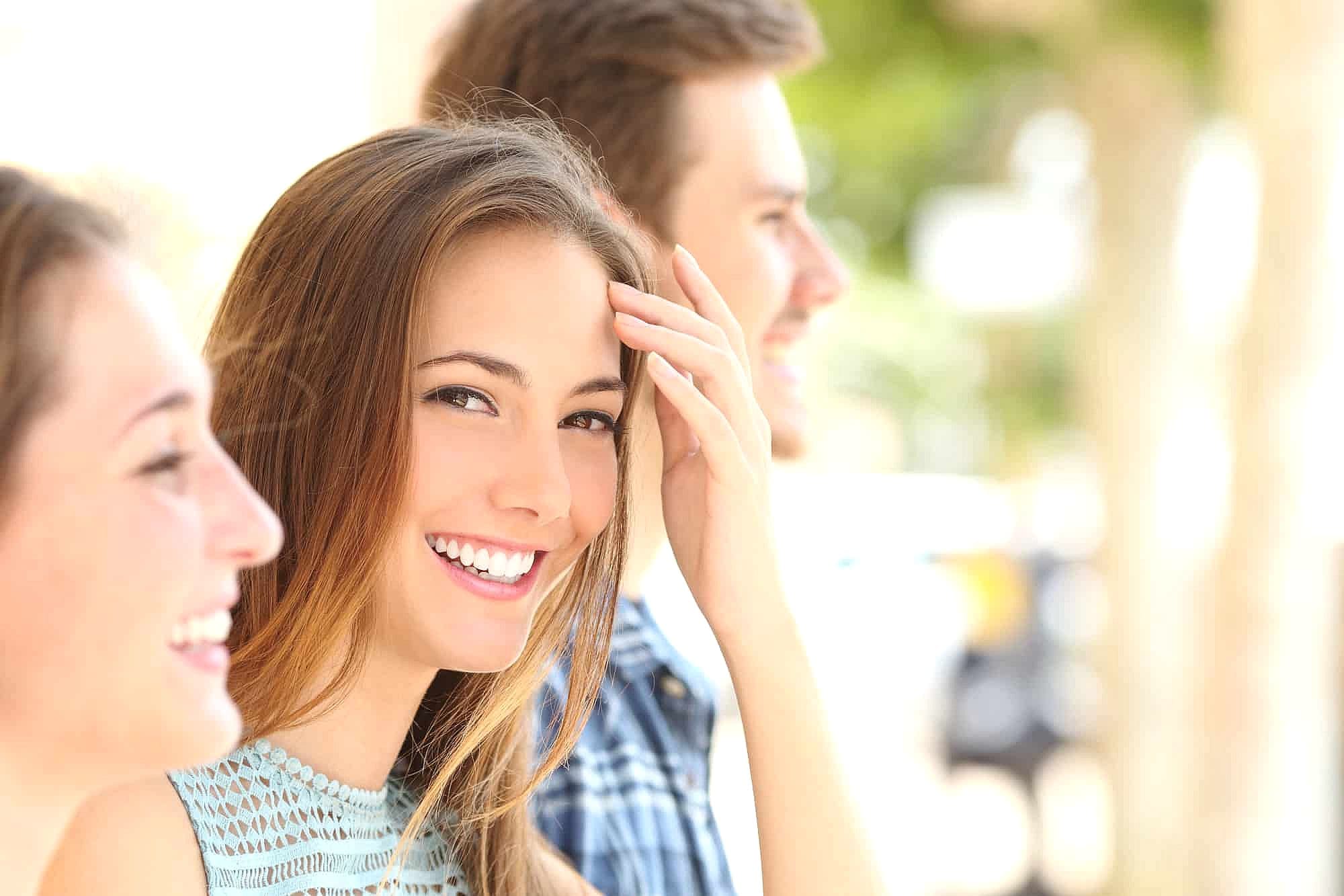 Three young adults enjoy a sunny day near Herbst Appliance, with the woman in front smiling at the camera while two friends beside her look away and smile. This cheerful scene captures the spirit of Smile City Orthodontics in Sunnyvale, TX.