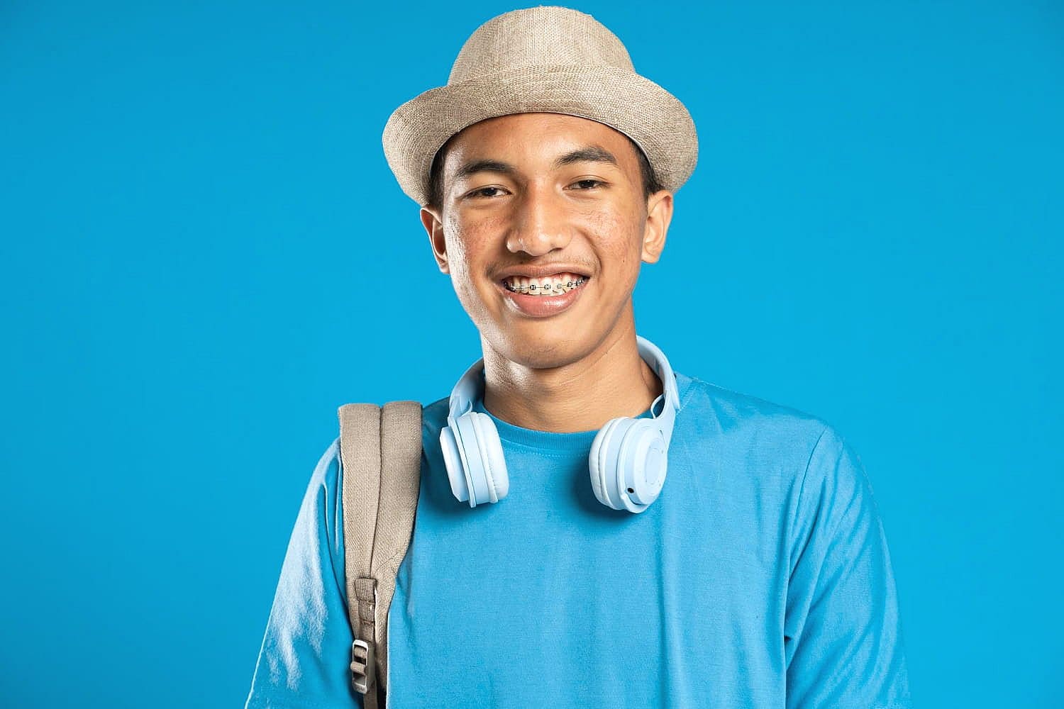 A young person in Sunnyvale, TX is pictured with an underbite, smiling with braces while wearing a hat, blue shirt, headphones around their neck, and a backpack strap over one shoulder against a blue background—representing Smile City Orthodontics.