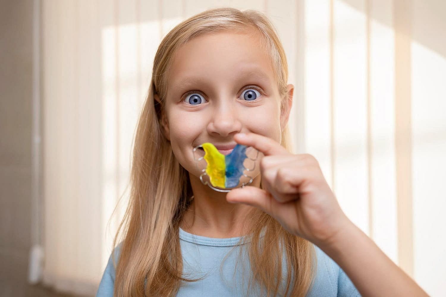 At Smile City Orthodontics in Sunnyvale, TX, a girl with long blonde hair holds a vibrant retainers featuring blue and yellow stripes, highlighting the creative options offered by their Children’s Orthodontist.