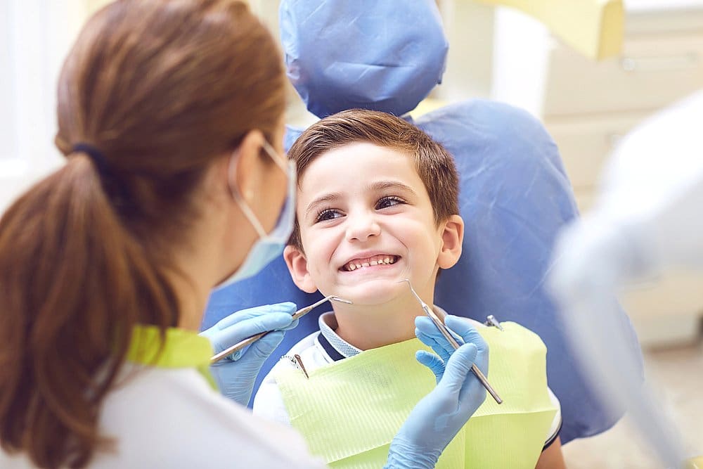 At Smile City Orthodontics in Sunnyvale TX, a children’s orthodontist examines a smiling young boy’s teeth during a checkup, with both wearing protective gear in the dental office.