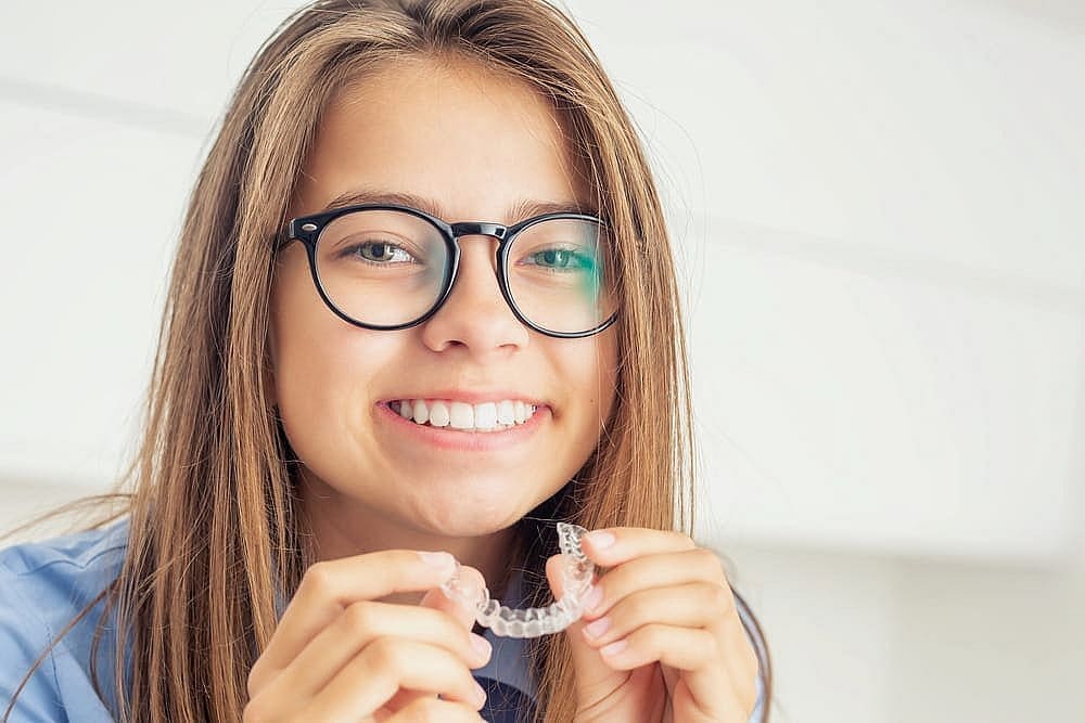 A young woman with long hair and glasses smiles while holding clear aligners, highlighting the modern solutions offered by Smile City Orthodontics in Sunnyvale, TX, beyond traditional children's orthodontics.