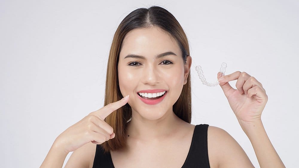 A woman smiles and points to her teeth while holding a clear dental aligner, highlighting the Invisalign payment plan benefits at Smile City Orthodontics in Sunnyvale TX, against a plain white background.
