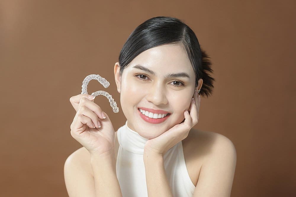 A woman smiles as she holds two clear dental aligners in her left hand against a plain brown background, demonstrating the benefits of an Invisalign payment plan at Smile City Orthodontics in Sunnyvale, TX.