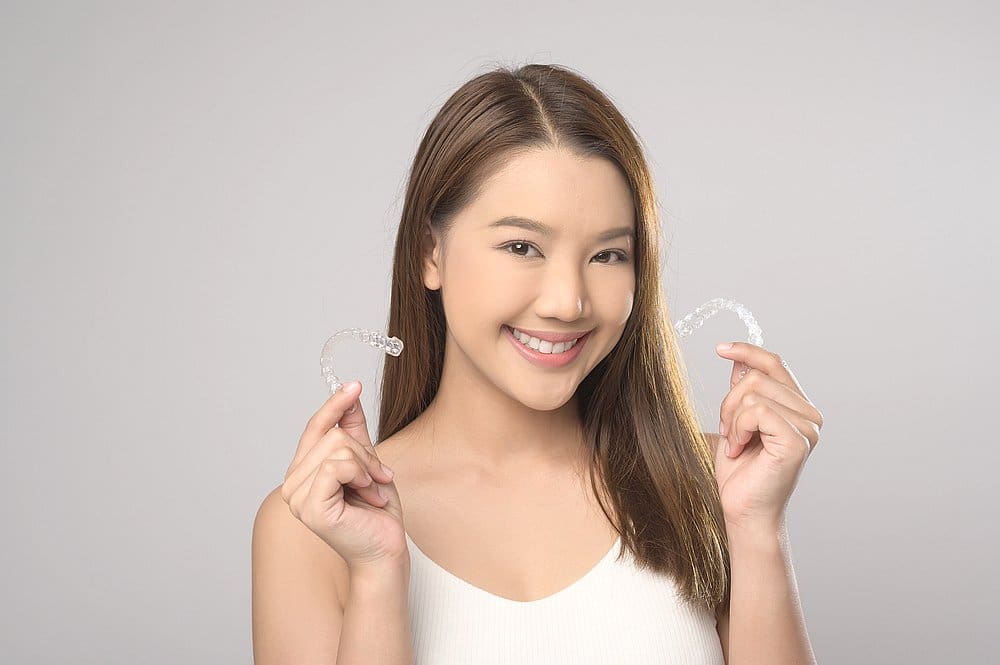 A woman with straight brown hair smiles and holds clear dental aligners against a plain background, showcasing the benefits of Invisalign at Smile City Orthodontics in Sunnyvale, TX—ideal for those exploring Invisalign insurance options.