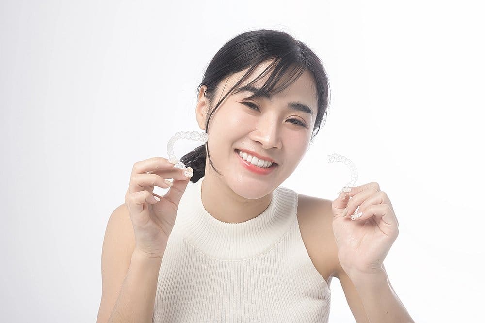 A woman in a white sleeveless top smiles and displays two clear dental aligners against a plain white background, highlighting the Invisalign payment plan offered by Smile City Orthodontics in Sunnyvale, TX.