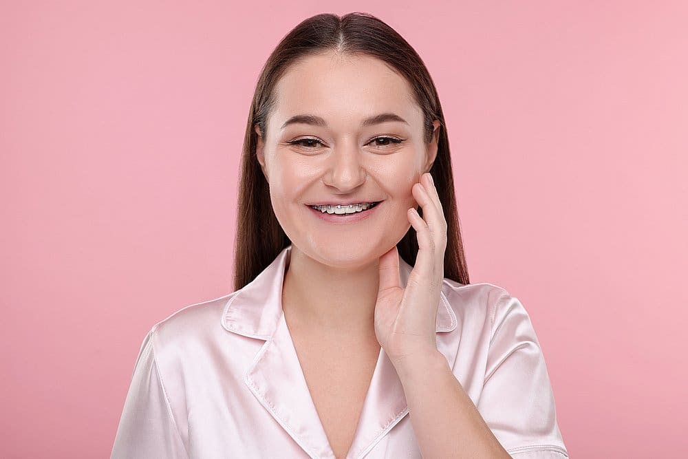 Showcasing the confidence that comes with adult braces, a woman with straight brown hair and braces smiles and touches her cheek in front of a pink background. This image highlights the transformative results provided by Smile City Orthodontics in Sunnyvale, TX.