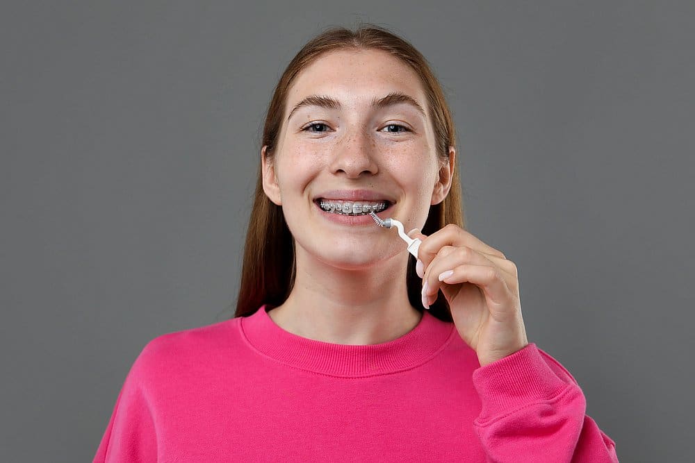 At Smile City Orthodontics in Sunnyvale TX, a young person with adult braces demonstrates proper oral care by using an interdental brush, dressed in a bright pink sweatshirt against a simple gray background.