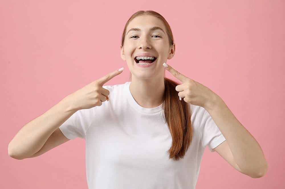 A young woman with long red hair wearing a white t-shirt smiles and points to her metal braces in a photo for Smile City Orthodontics in Sunnyvale TX, set against a pink background.