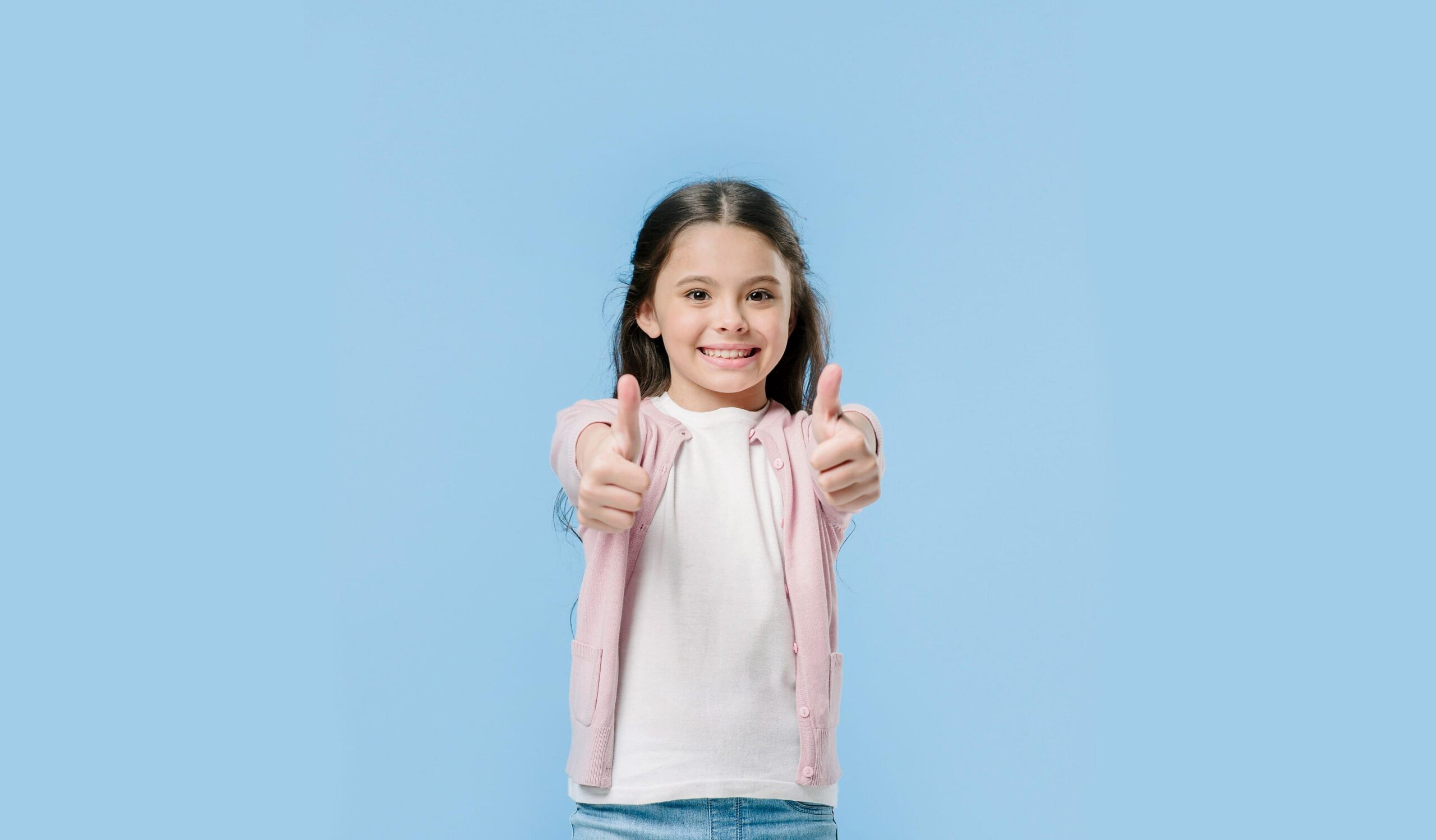 Beaming with confidence in front of a light blue background, a young girl in a white shirt and pink cardigan proudly shows off her affordable braces with two thumbs up—another happy patient at Smile City Orthodontics in Sunnyvale, TX.