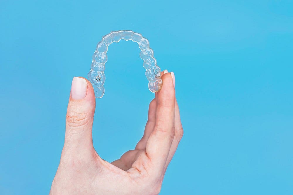 A hand holds a clear dental aligner against a plain blue background, showcasing options like Invisalign cost at Smile City Orthodontics in Sunnyvale, TX.