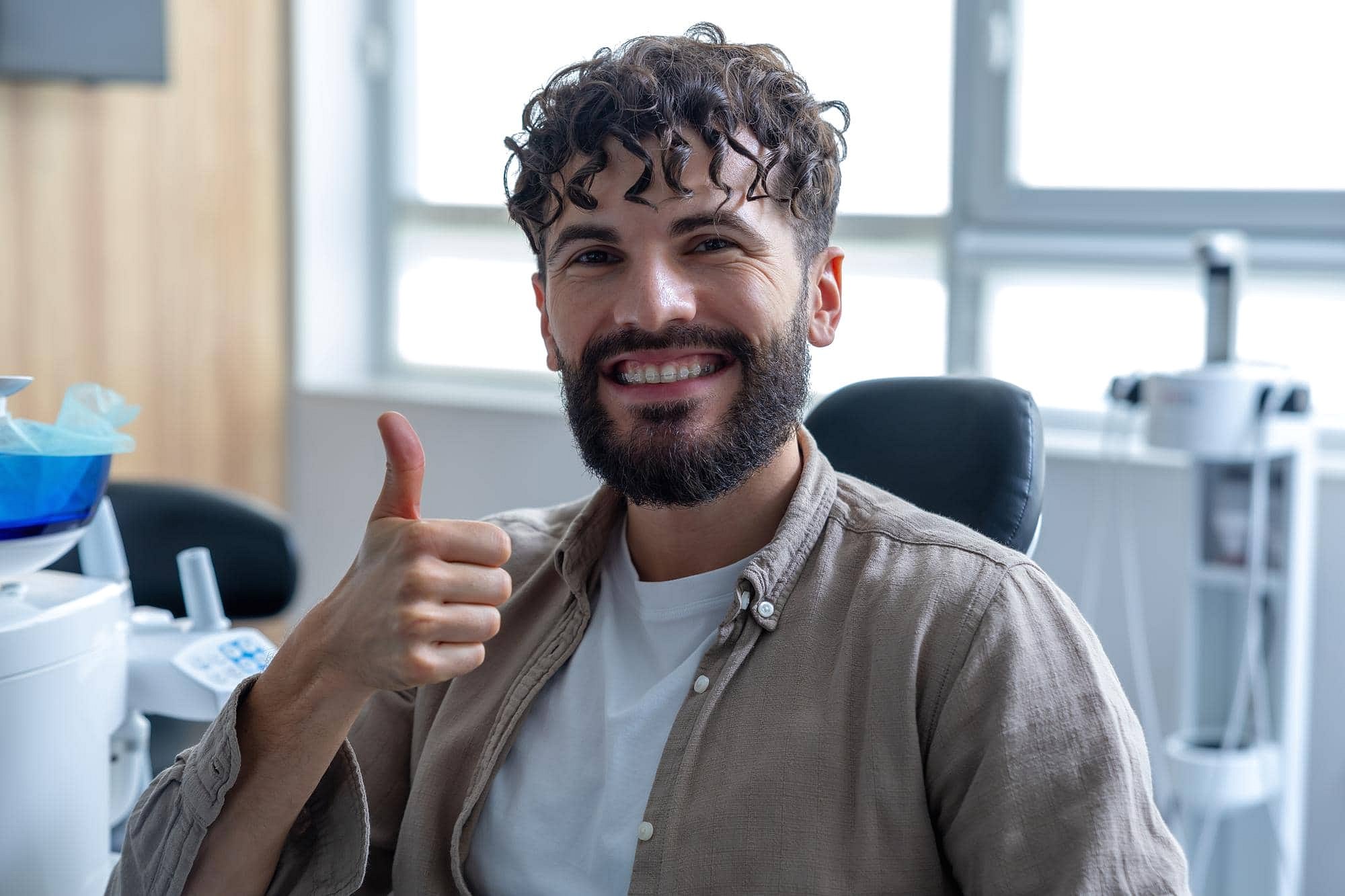 At Smile City Orthodontics in Sunnyvale TX, a man with curly hair and a beard smiles confidently indoors, showing off his ceramic braces and giving a thumbs-up.