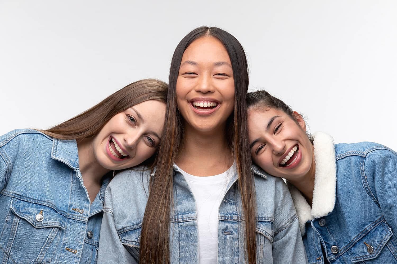 Three young women in denim jackets stand close together, smiling and laughing against a plain white background—the kind of confidence you can expect at Smile City Orthodontics in Sunnyvale, TX.