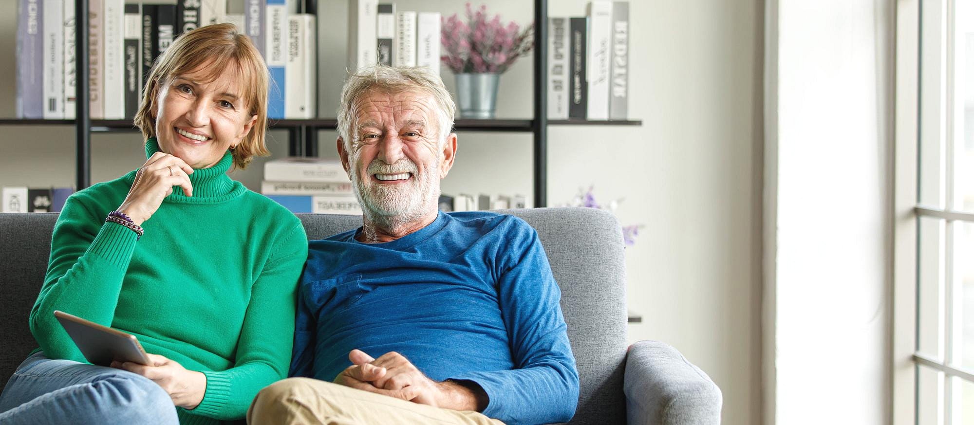 A smiling older couple sits together on a couch indoors, with the woman holding a tablet—possibly researching orthodontic insurance plans for Smile City Orthodontics in Sunnyvale, TX. Behind them, a bookshelf displays books and plants.