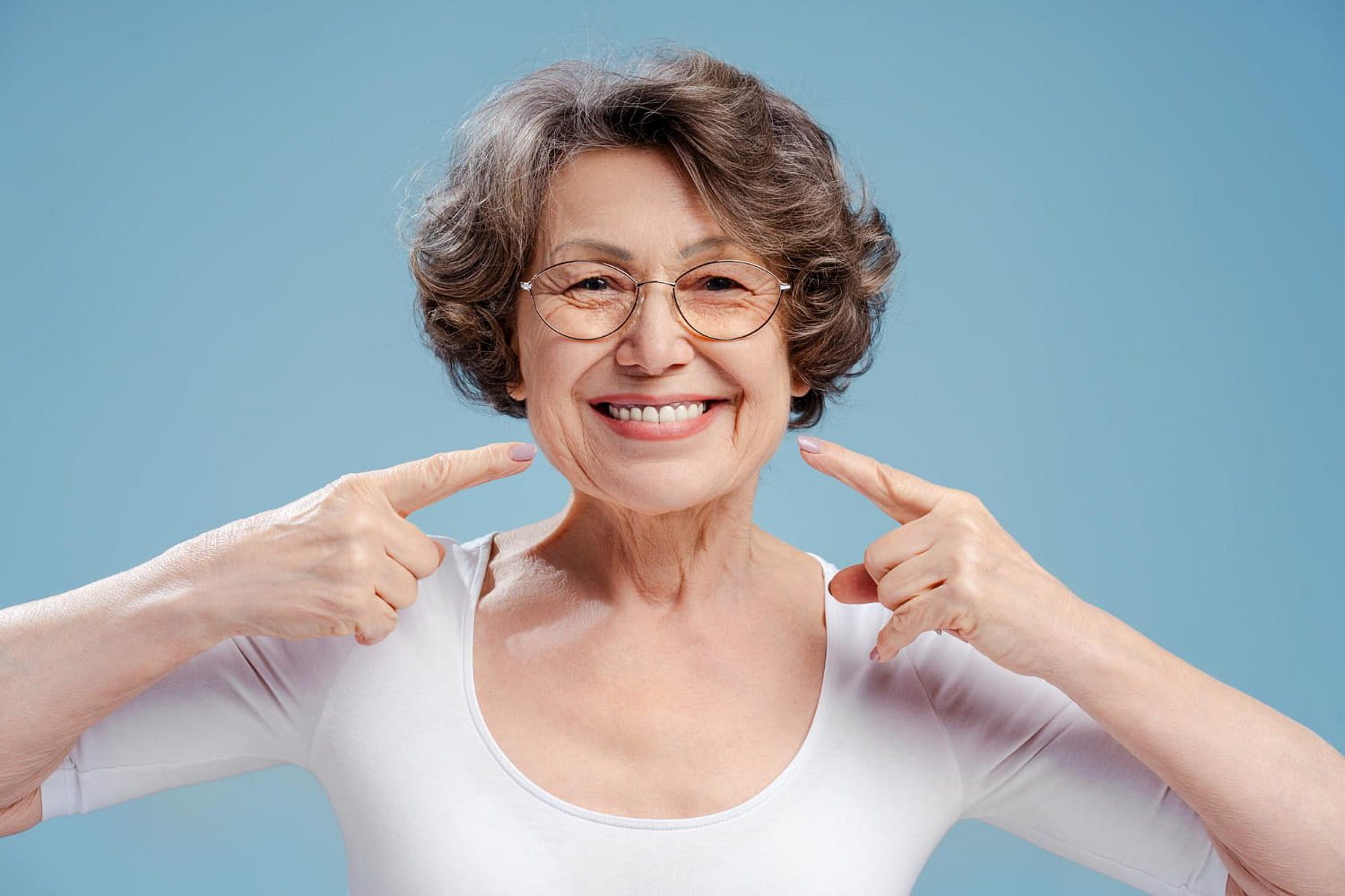 A smiling older woman with short brown hair and glasses, dressed in a white top, points to her teeth against a plain blue background, showcasing the benefits of Orthodontic Insurance Plans at Smile City Orthodontics in Sunnyvale, TX.