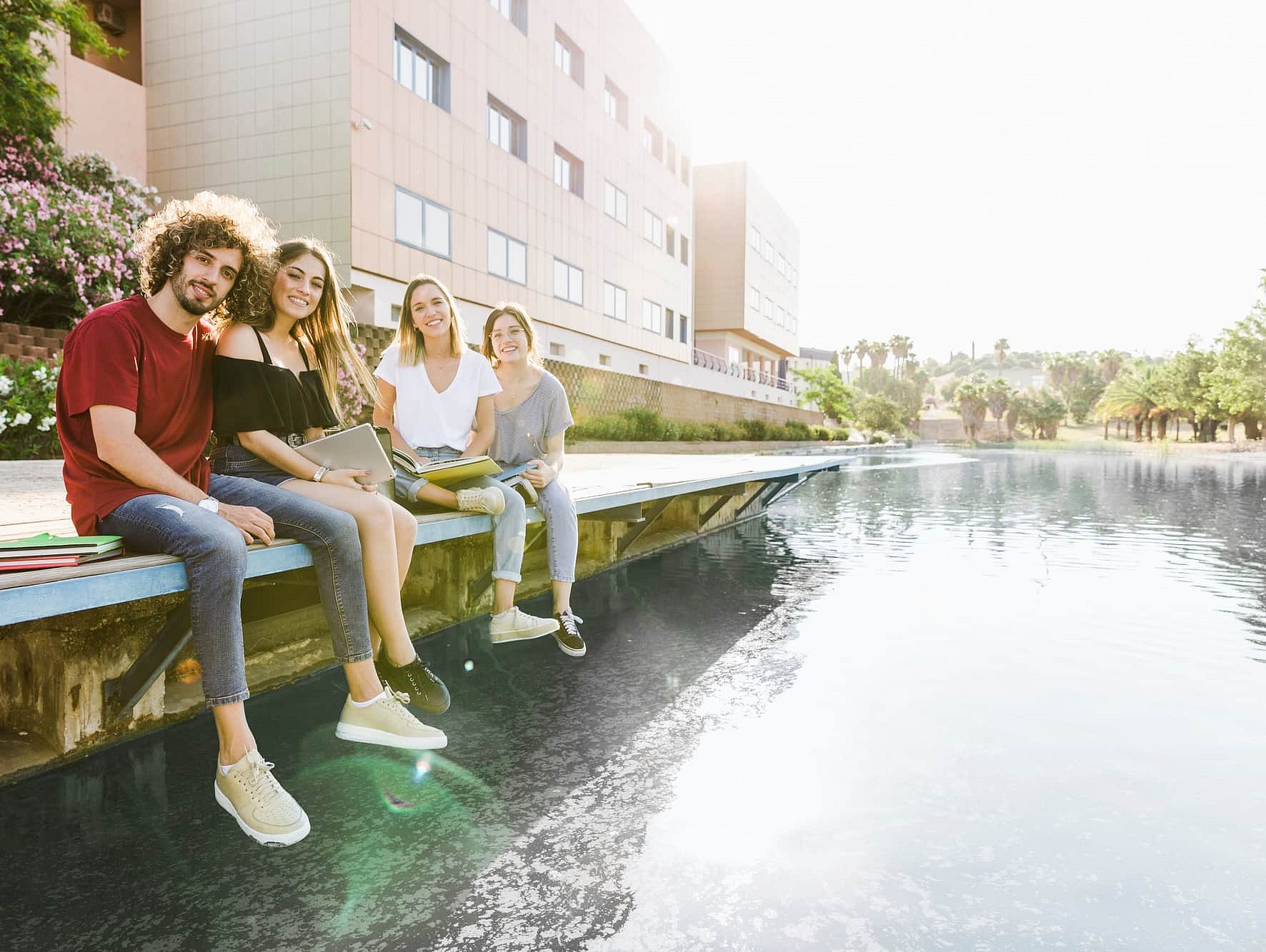 By a pool outside a modern building in Sunnyvale TX, four young adults braces relax and smile, holding notebooks and a laptop—one proudly displaying their adult braces from Smile City Orthodontics.