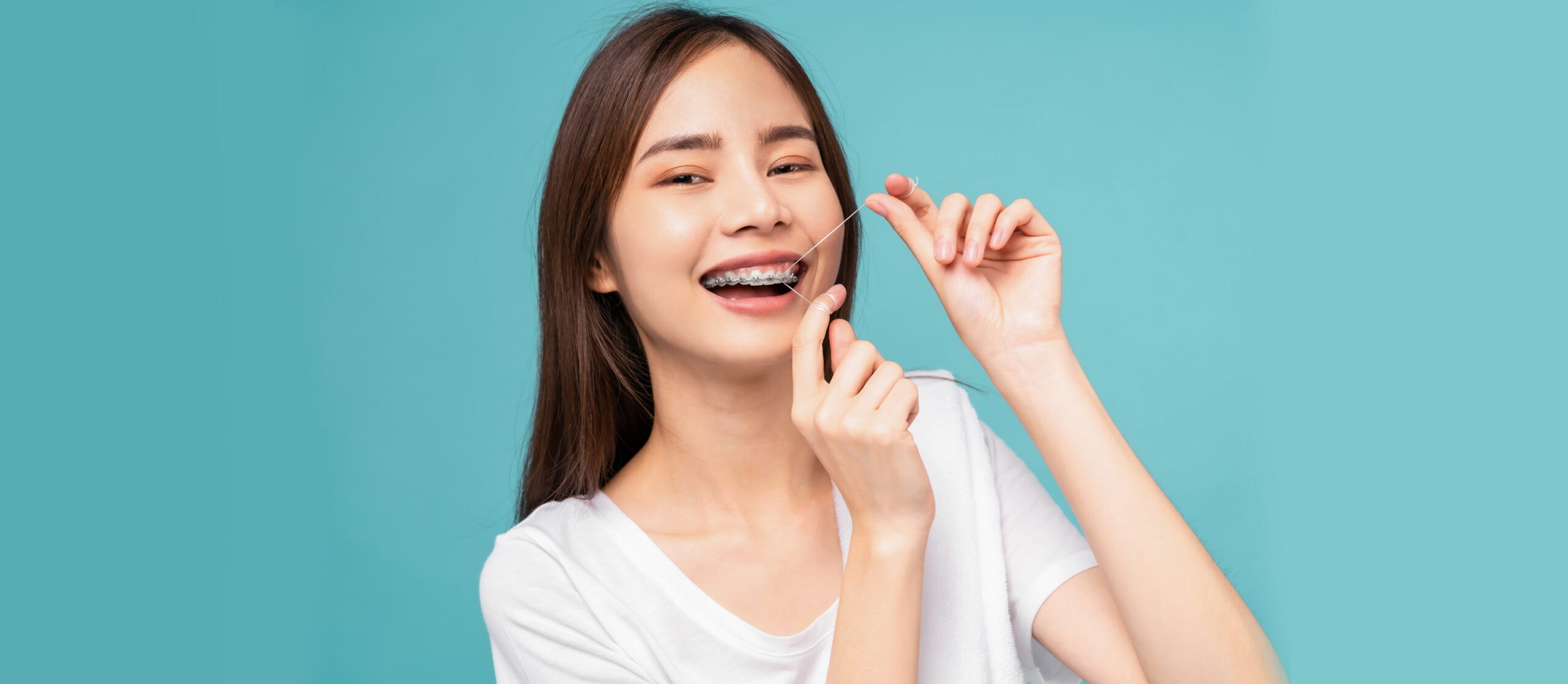 A young woman with long hair and braces smiles while flossing her teeth against a plain blue background, showcasing the benefits of adult orthodontics at Smile City Orthodontics in Sunnyvale, TX.