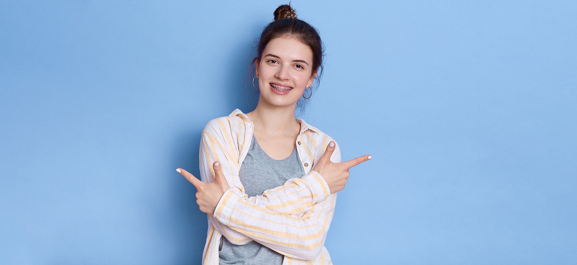 A young person in Sunnyvale, TX points both index fingers to the sides, flashing a bright smile with braces that showcase the value of braces cost at Smile City Orthodontics, all set against a plain blue background.