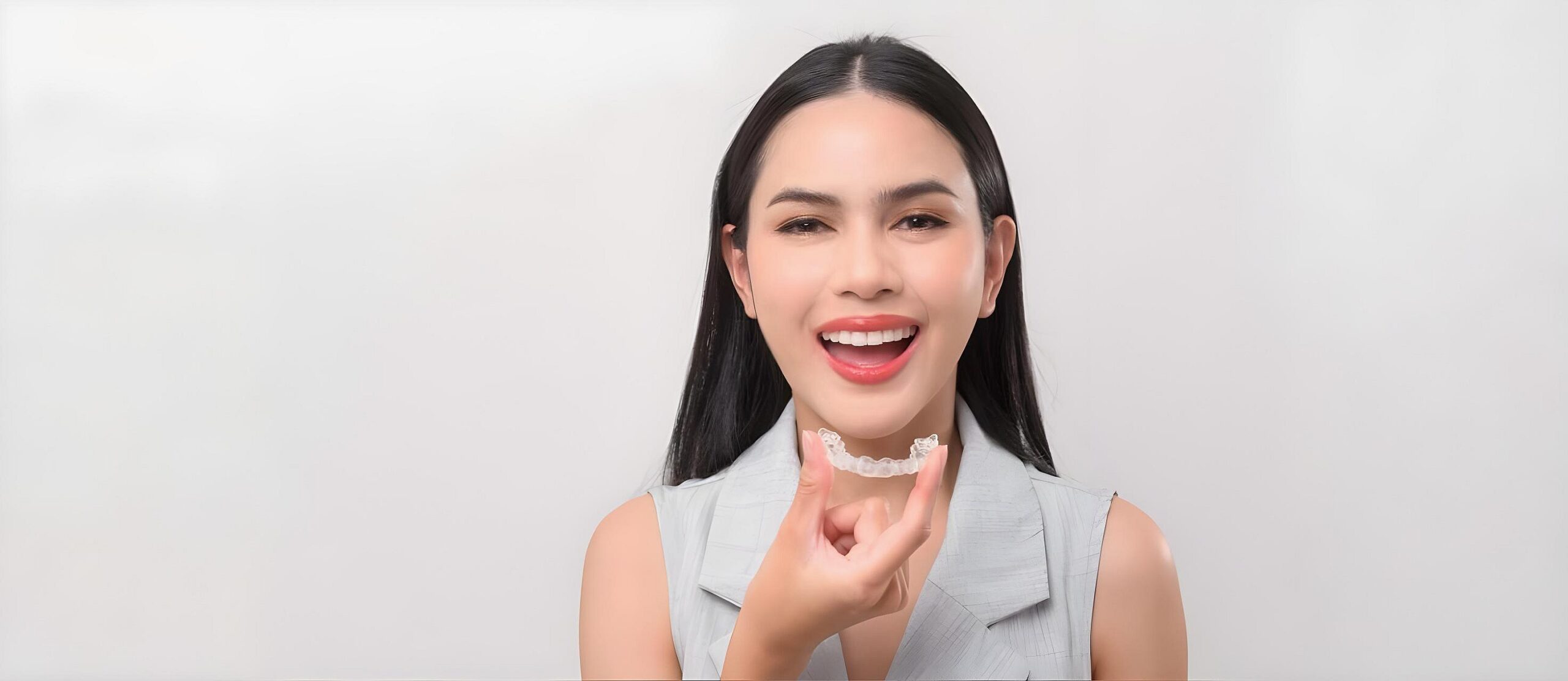 A woman smiles while holding a clear aligner near her mouth, showcasing the Invisalign vs Braces choice at Smile City Orthodontics in Sunnyvale, TX.