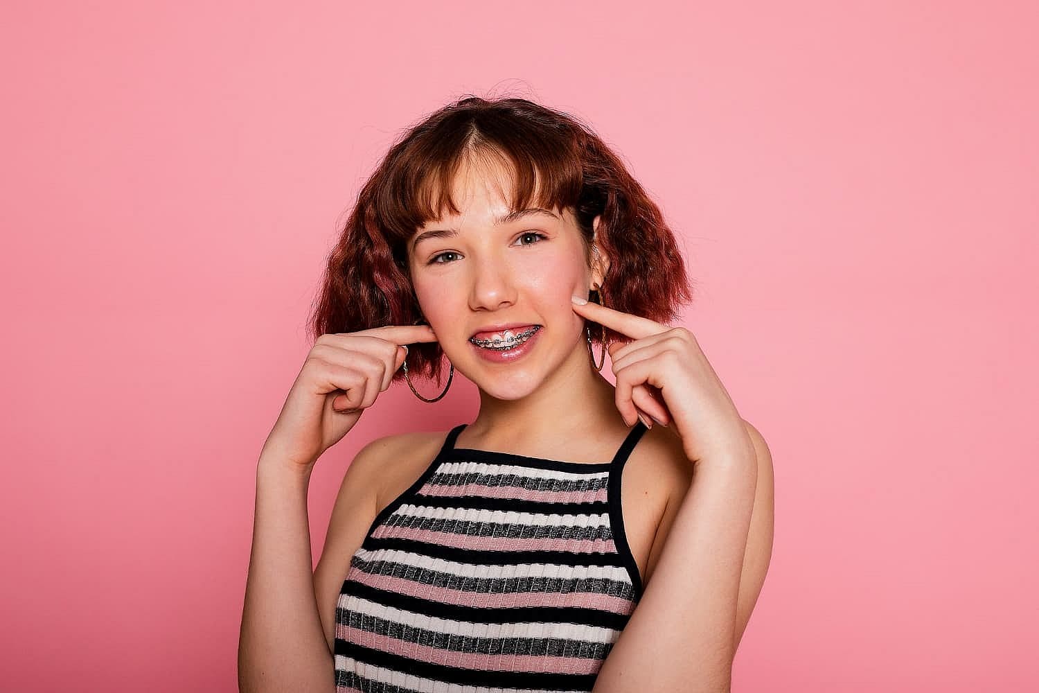 Against a plain pink background, a teenager with short brown hair and braces smiles and points to her cheeks, embodying the essence of Invisalign vs Braces at Smile City Orthodontics in Sunnyvale, TX.