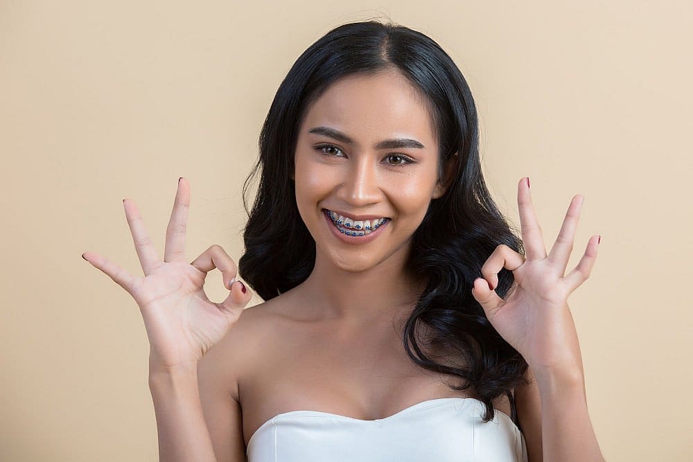 Confident after getting metal adult braces at Smile City Orthodontics in Sunnyvale TX, a woman with long black hair smiles and flashes "OK" hand signs against a plain beige background.
