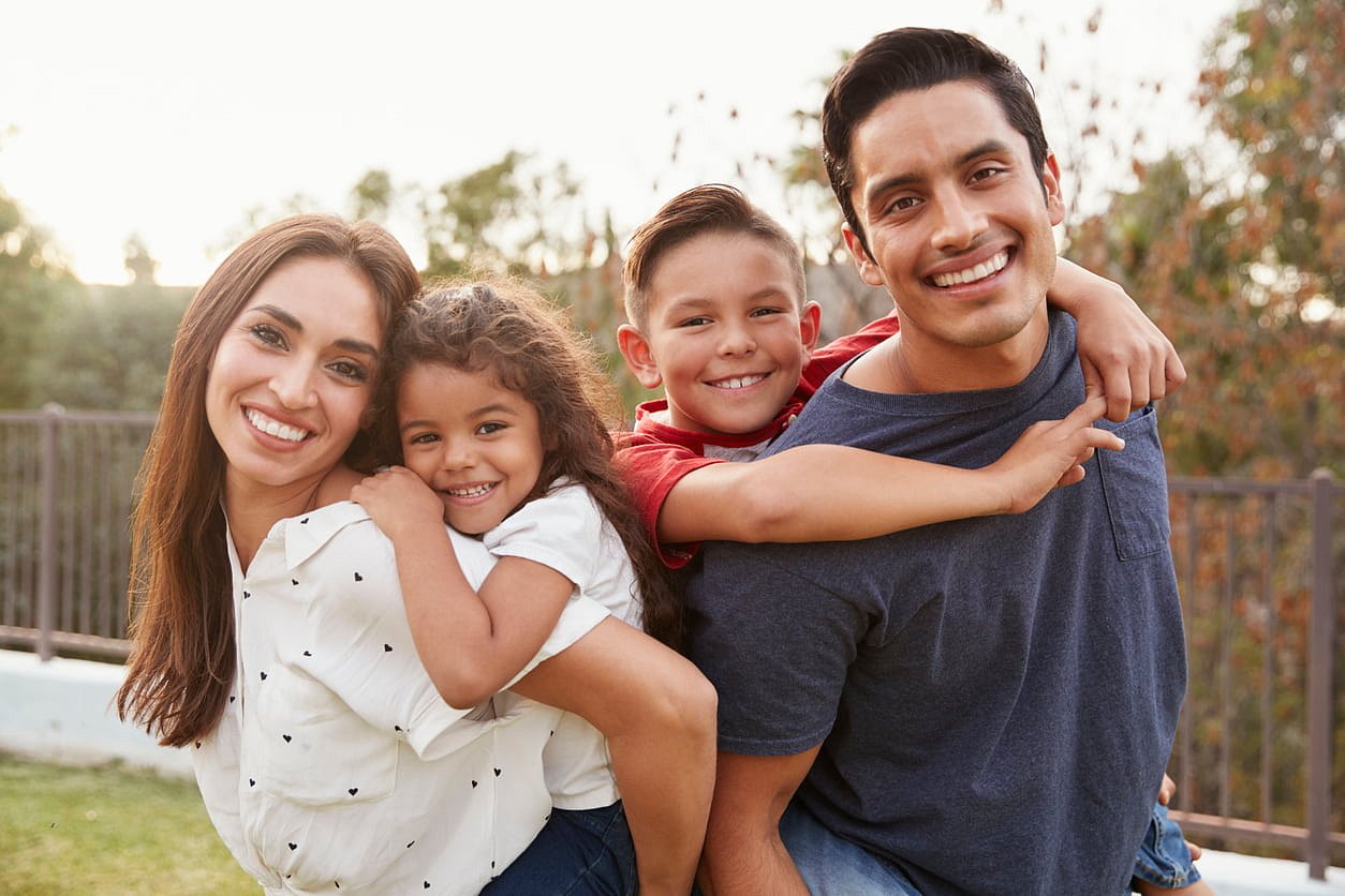 A smiling woman and man give piggyback rides to a young girl and boy, all showing off their healthy smiles thanks to affordable braces from Smile City Orthodontics in Sunnyvale, TX.