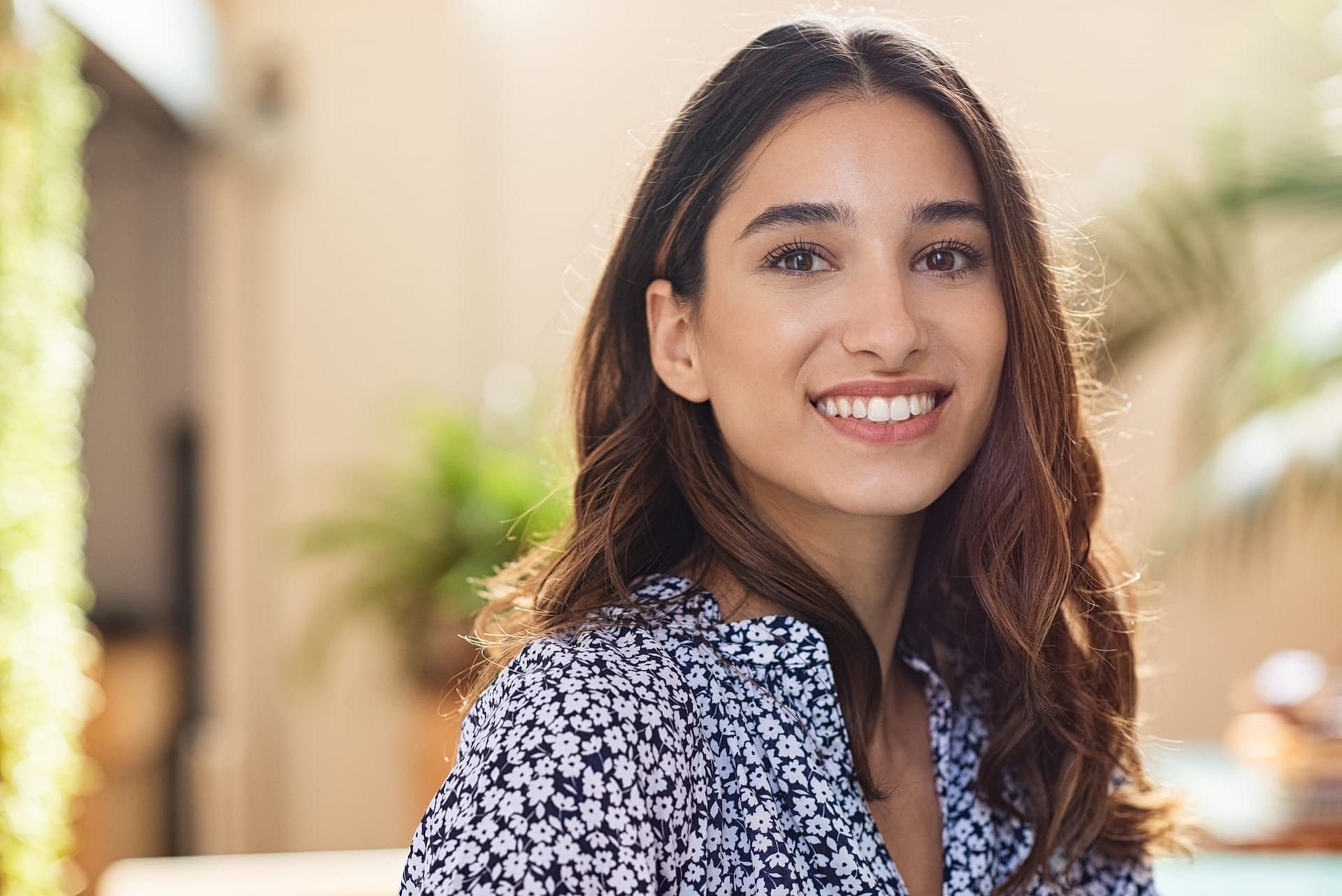 In a bright indoor setting at Smile City Orthodontics in Sunnyvale TX, a woman with long brown hair and a floral shirt smiles confidently at the camera, reflecting the assurance that comes from orthodontic insurance coverage amid blurred plants in the background.