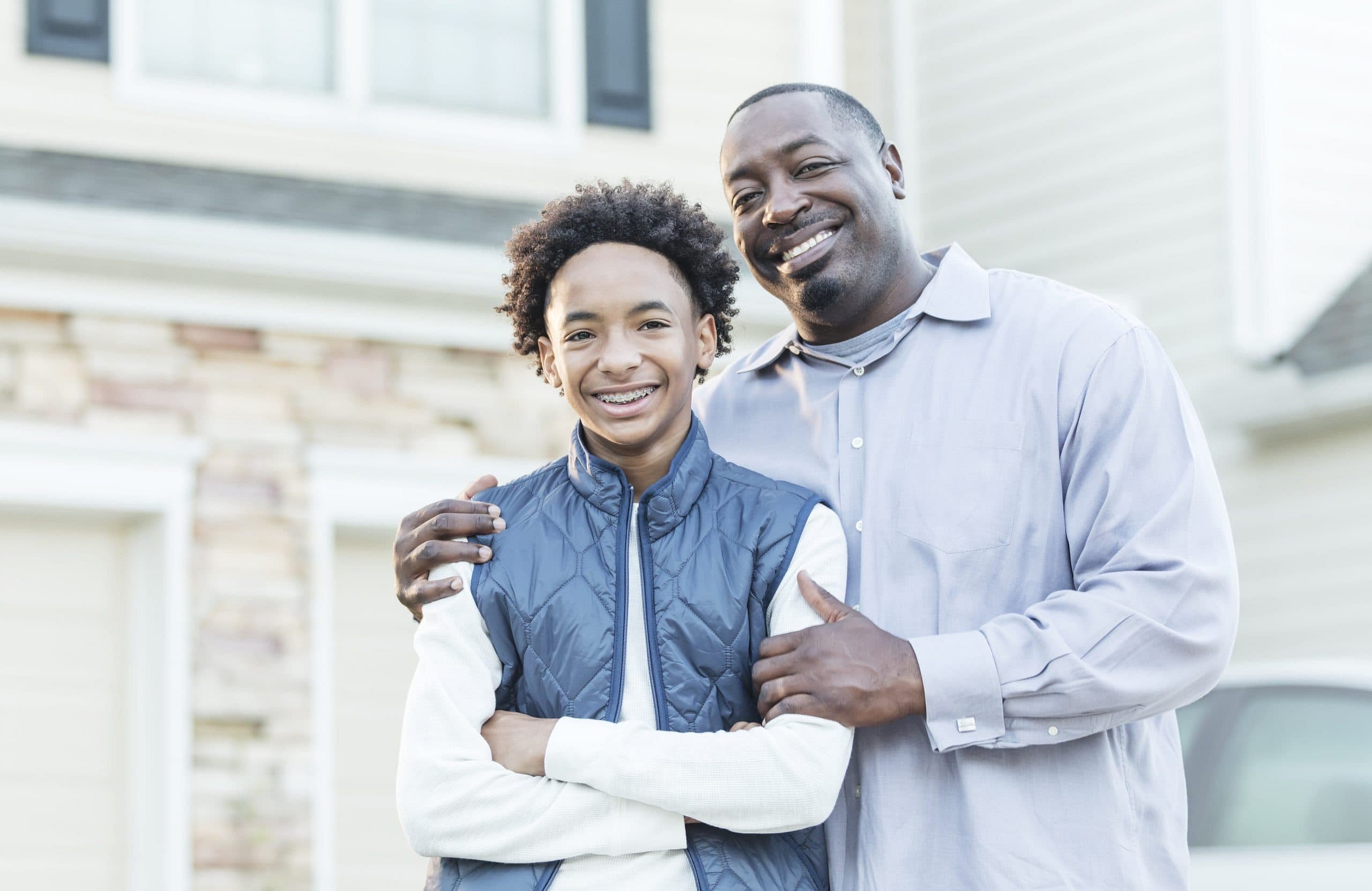 In Sunnyvale, TX, a man—confident in his smile thanks to adult orthodontics at Smile City Orthodontics—stands outside a house with a boy in a blue vest, both smiling at the camera.