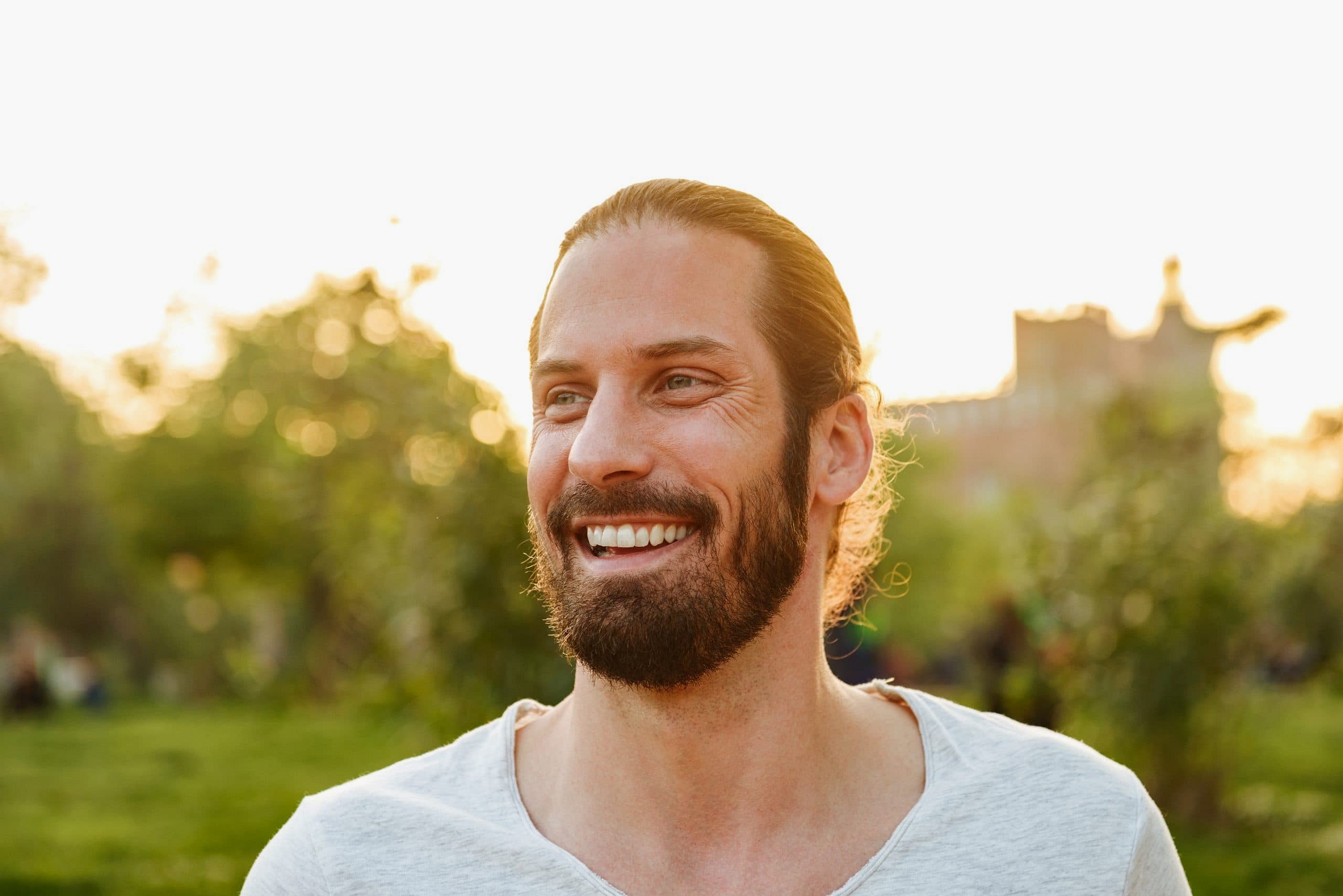 Outdoors in a park in Sunnyvale TX, a bearded man with long hair tied back smiles, his overbite subtly visible against greenery and a blurred building—an everyday scene Smile City Orthodontics understands well.