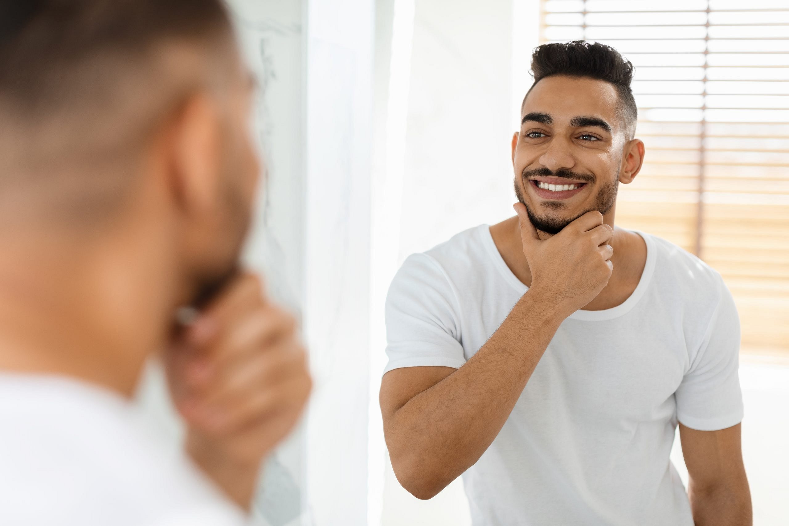 A man with short dark hair and a beard, wearing a white t-shirt, admires his new smile in the mirror after jaw surgery at Smile City Orthodontics in Sunnyvale TX, standing in a bright room.