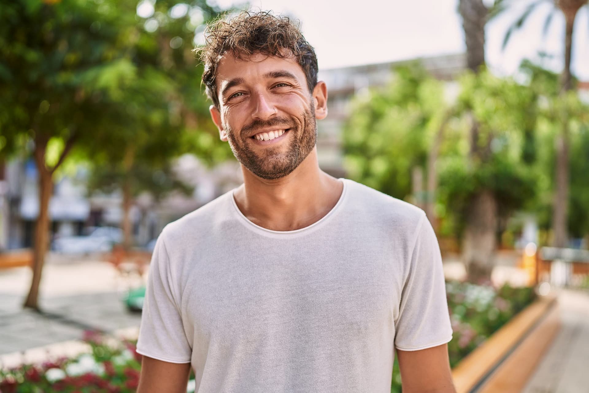 Outdoors in Sunnyvale TX, a man with curly hair, a beard, and metal braces smiles in front of trees and greenery while wearing a white t-shirt—capturing the confident results from Smile City Orthodontics.