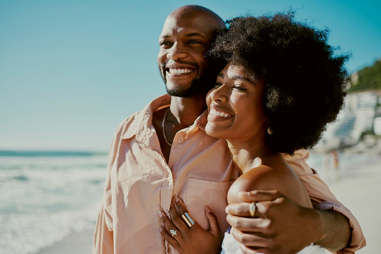 A couple on a sunny beach in Sunnyvale TX, smiles and embraces while displaying their Herbst Appliance, with the ocean and buildings behind them—capturing a happy moment at Smile City Orthodontics.