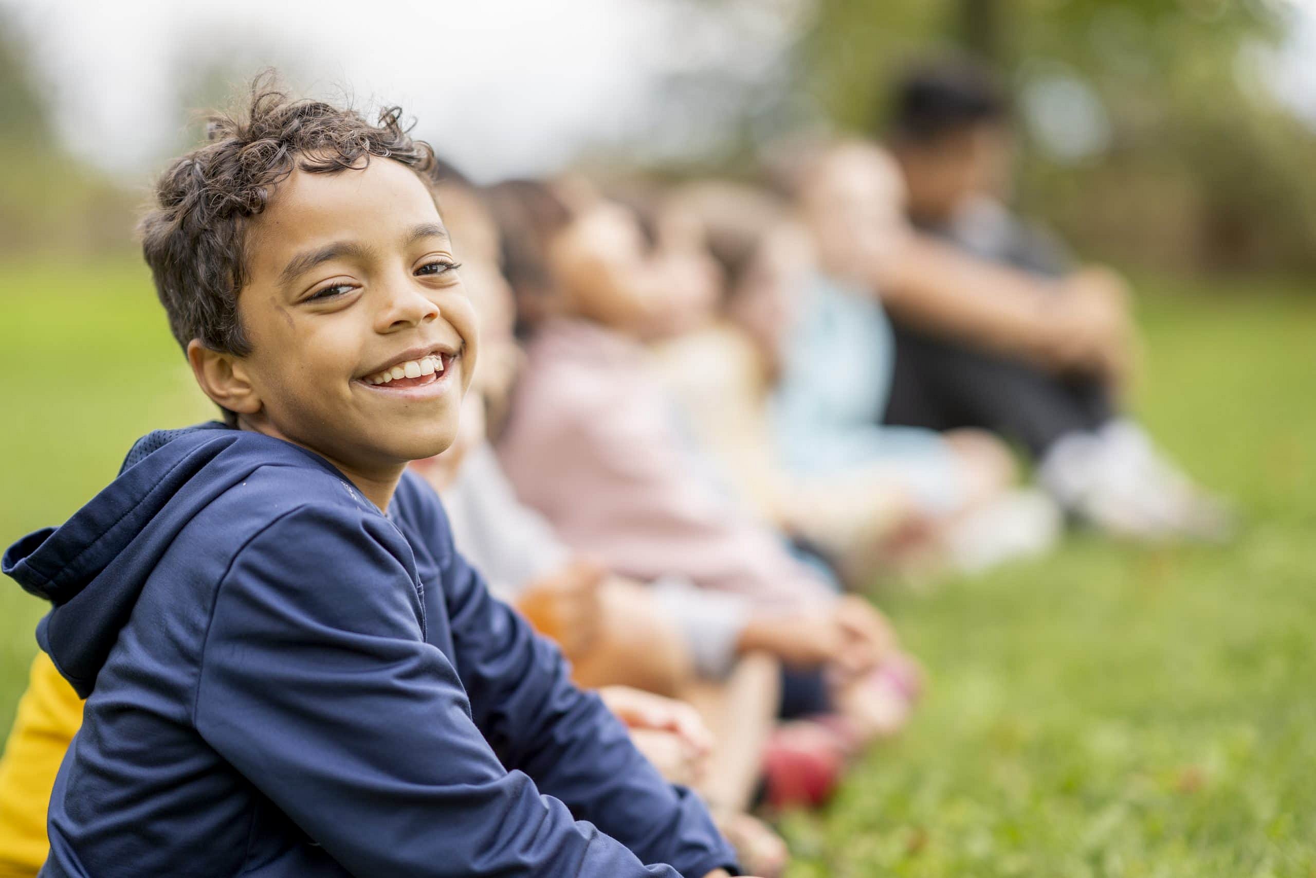 At Smile City Orthodontics in Sunnyvale TX, a boy in a navy hoodie displays a cheerful overbite while sitting on the grass and smiling at the camera, with other children blurred in the background.