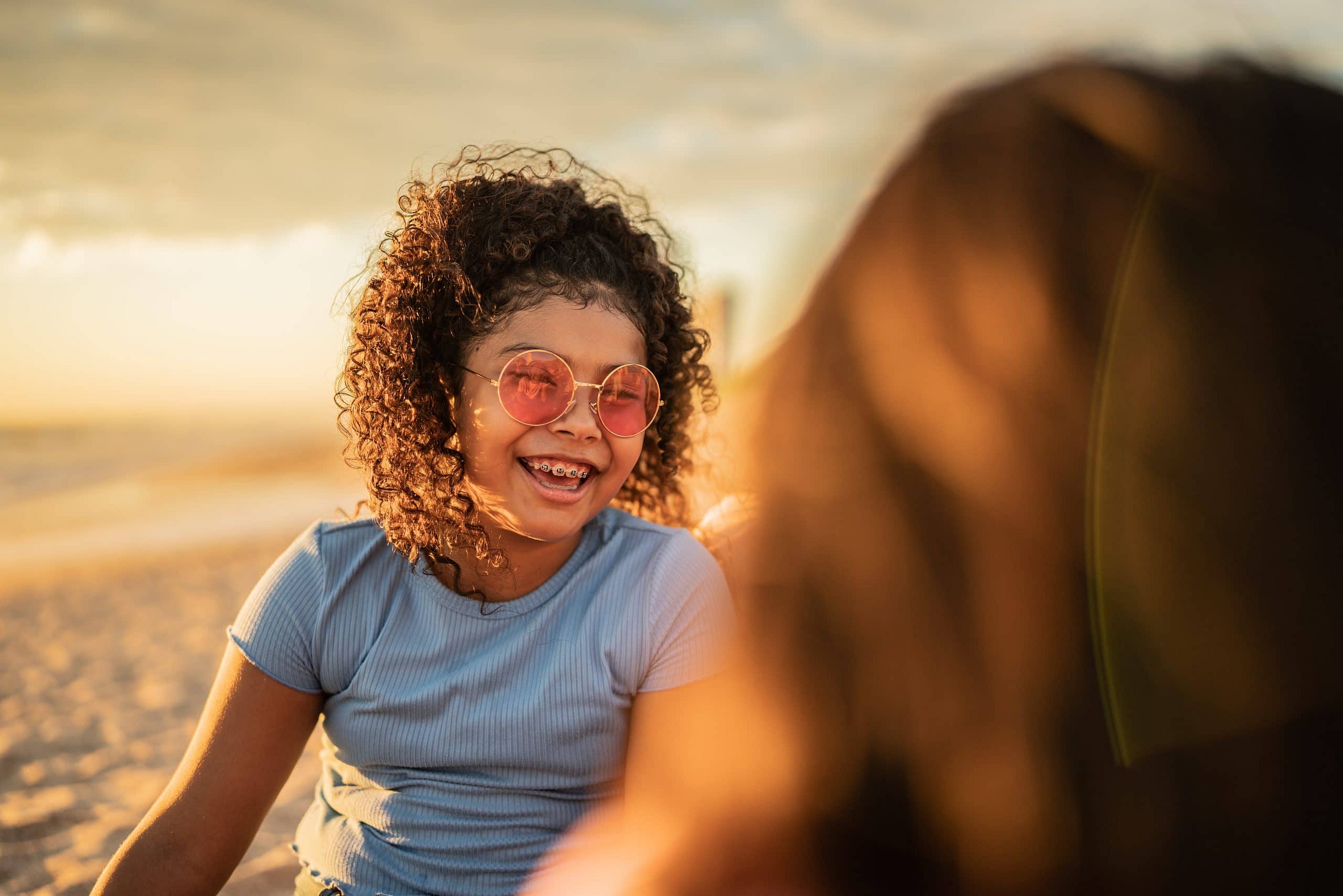 At Smile City Orthodontics in Sunnyvale TX, a girl with curly hair and pink sunglasses showing a slight underbite smiles on a sandy beach at sunset, with another person partially visible in the foreground.