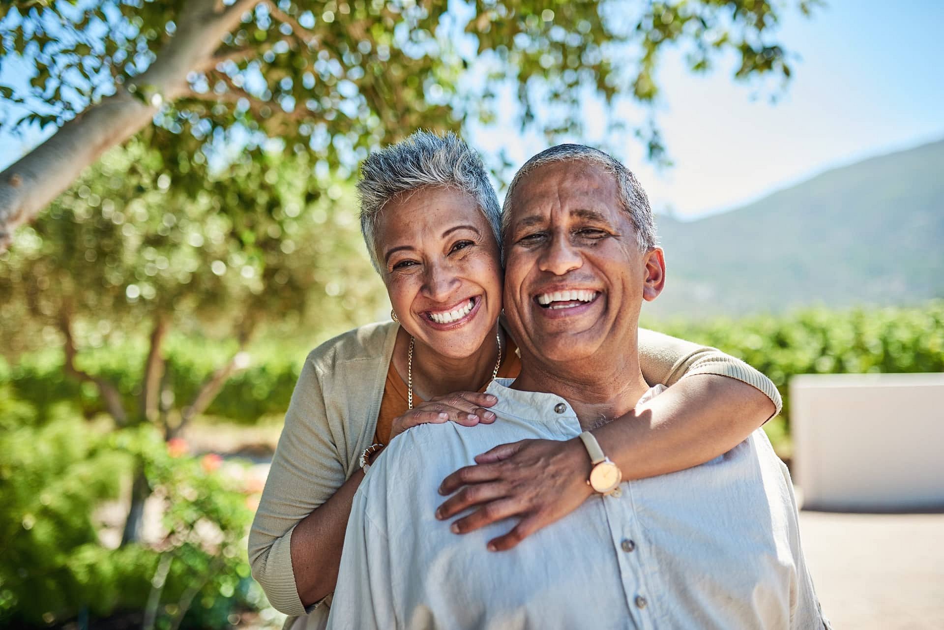 Smiling outdoors on a sunny day surrounded by lush greenery, an older couple embraces and enjoys each other's company—showcasing the healthy smiles made possible by Smile City Orthodontics in Sunnyvale, TX.