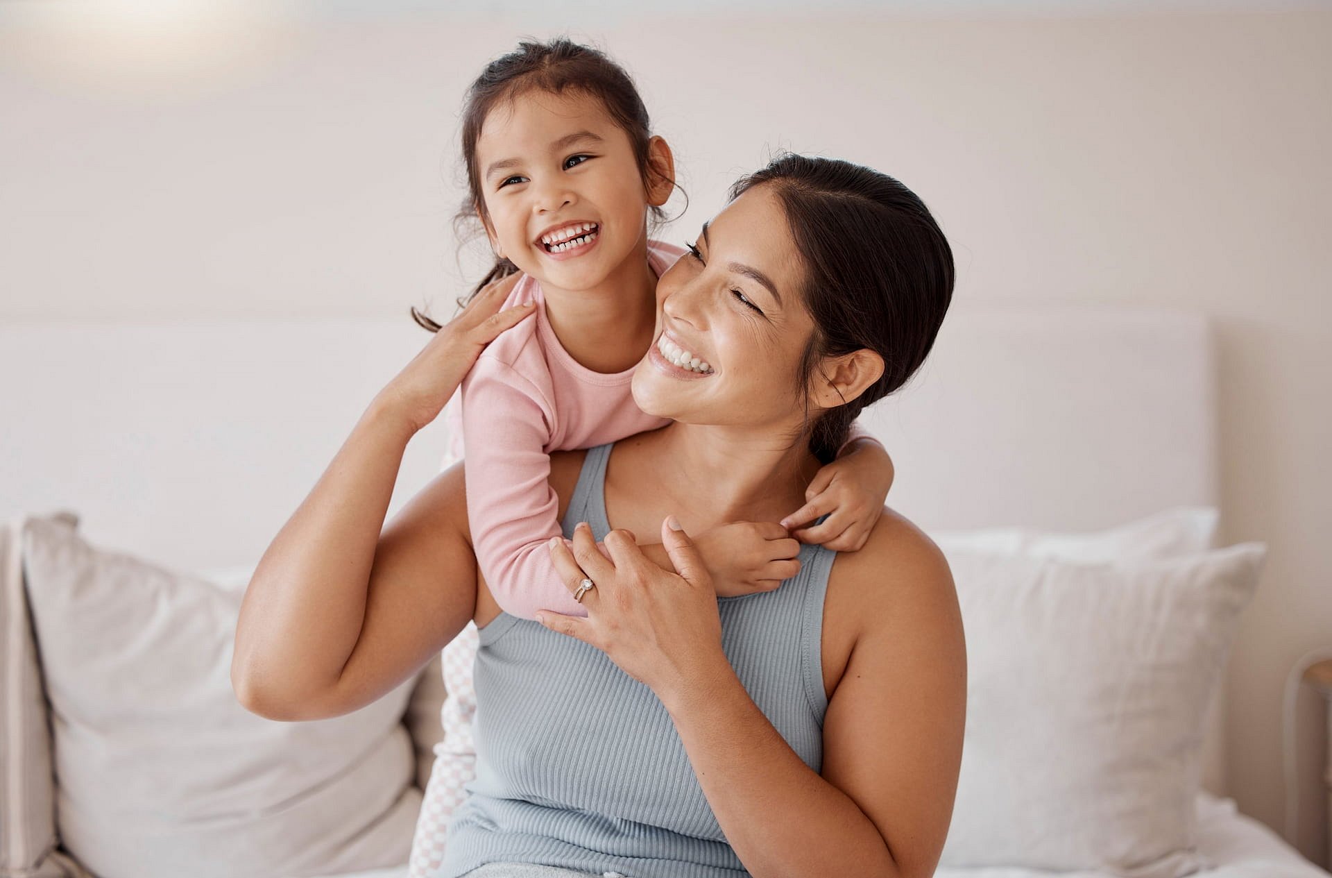 At Smile City Orthodontics in Sunnyvale TX, a woman sits on a bed, smiling as a young girl with metal braces hugs her from behind; both look happy and gaze in the same direction.