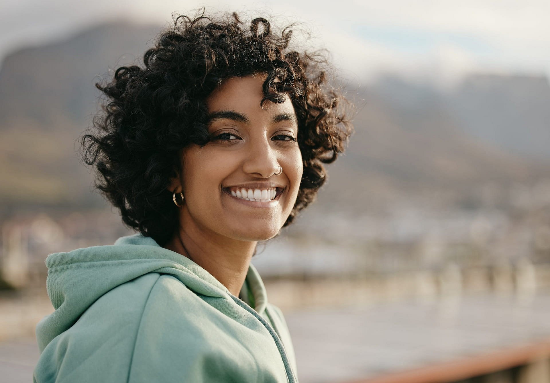 At Smile City Orthodontics in Sunnyvale TX, a young woman with short curly hair and braces smiles outdoors, dressed in a green hoodie with mountains and sky softly blurred behind her.