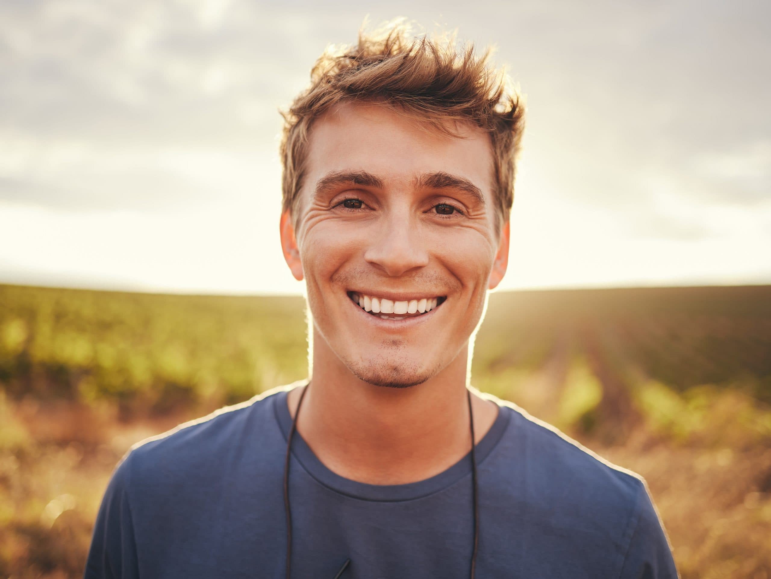 In a sunlit field in Sunnyvale TX, a young man with short brown hair, wearing a blue shirt and a cord around his neck, smiles at the camera with the relaxed expression typical of a mouth breather—capturing the welcoming spirit of Smile City Orthodontics.