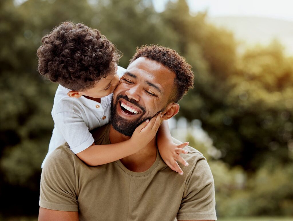 At Smile City Orthodontics in Sunnyvale TX, a smiling man is hugged and kissed on the cheek by a young child outdoors, with trees softly blurred in the background.