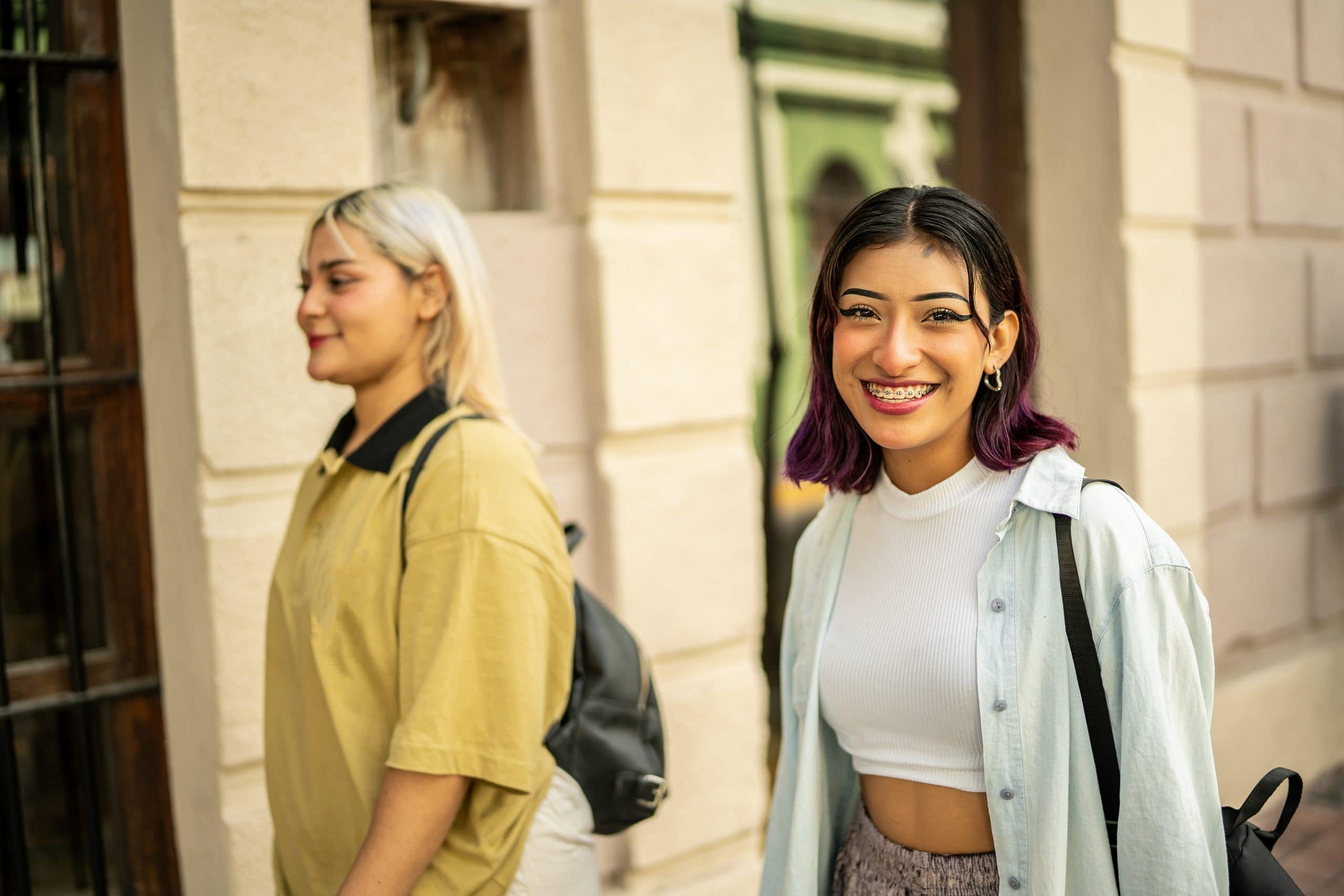 Outside Smile City Orthodontics in Sunnyvale TX, two young women with backpacks walk together—one in a yellow shirt looking ahead, while the other, a cheerful mouth breather in a white shirt, smiles at the camera.