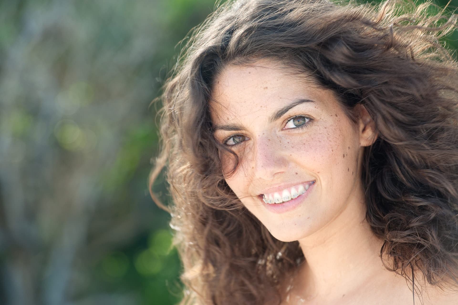 A young woman with curly brown hair and braces smiles outdoors, highlighting the benefits of adult orthodontics at Smile City Orthodontics in Sunnyvale, TX.