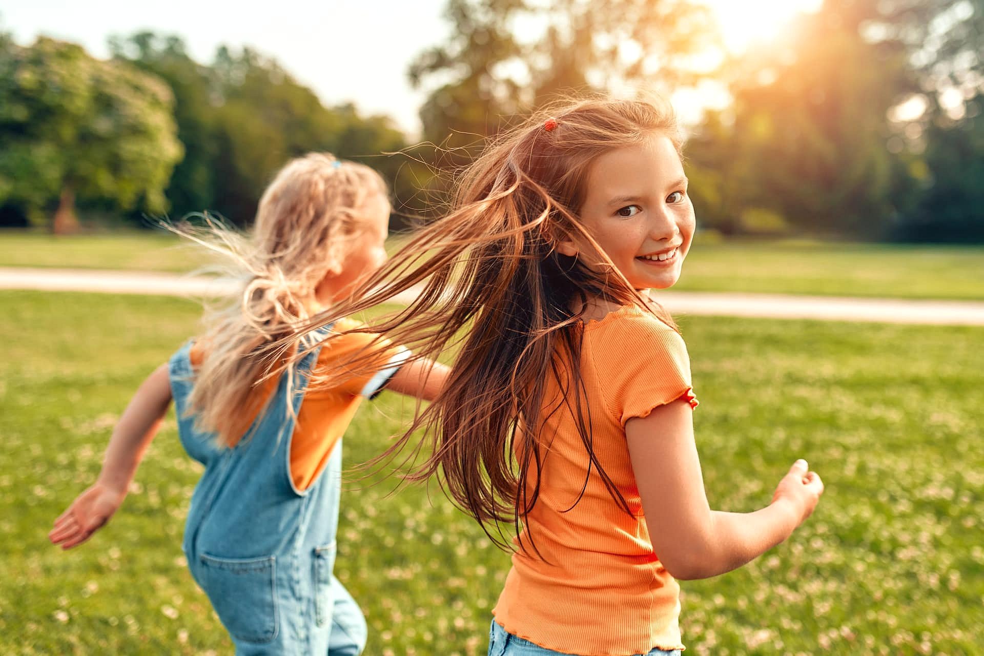 Confident smiles from a children’s orthodontist are on display as two young girls run and smile in a sunny, grassy park with trees in the background thanks to Smile City Orthodontics in Sunnyvale, TX.