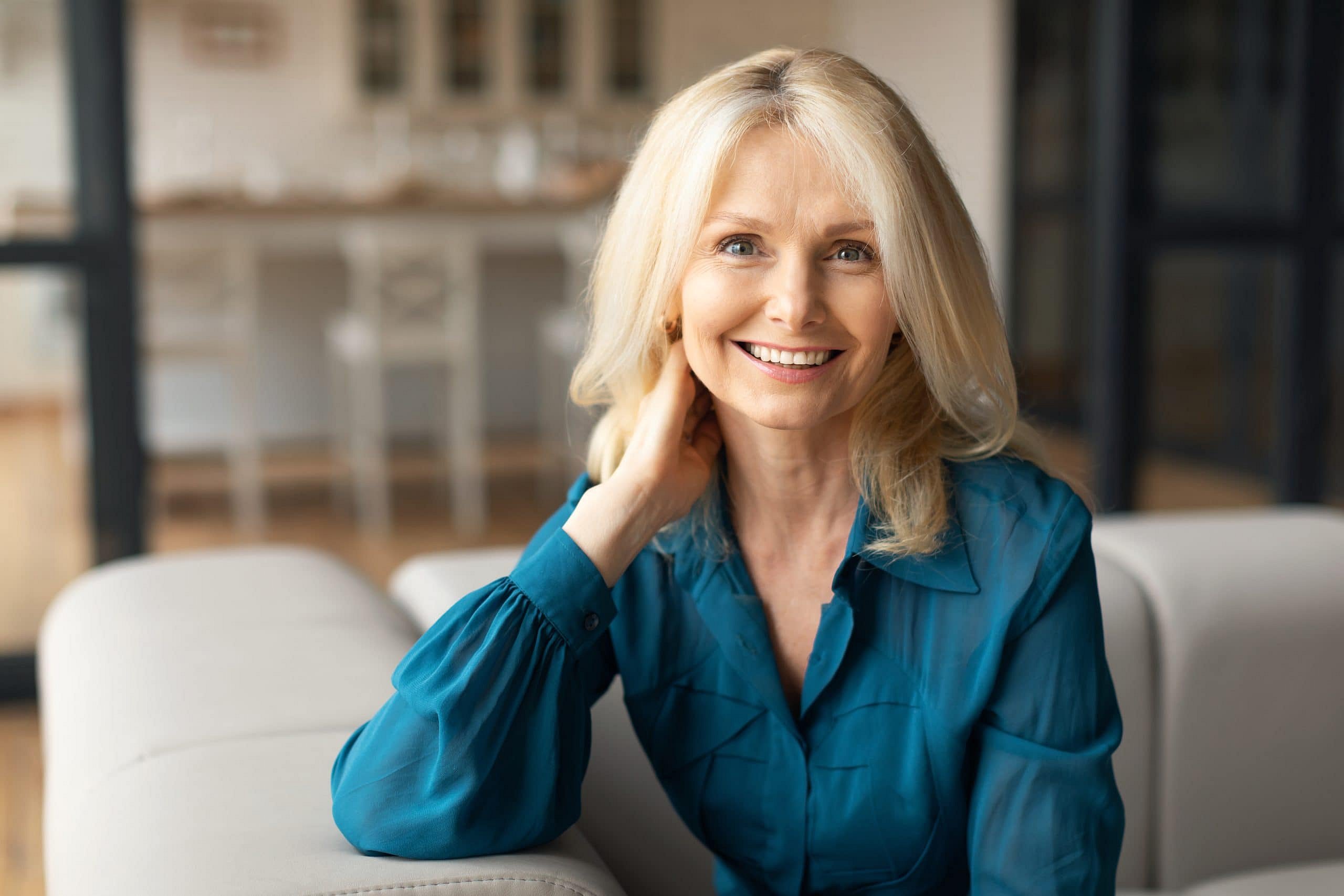 A middle-aged blonde woman in a teal blouse sits on a light sofa, smiling at the camera in her bright, modern living room while reviewing orthodontic insurance plans for Smile City Orthodontics in Sunnyvale, TX.
