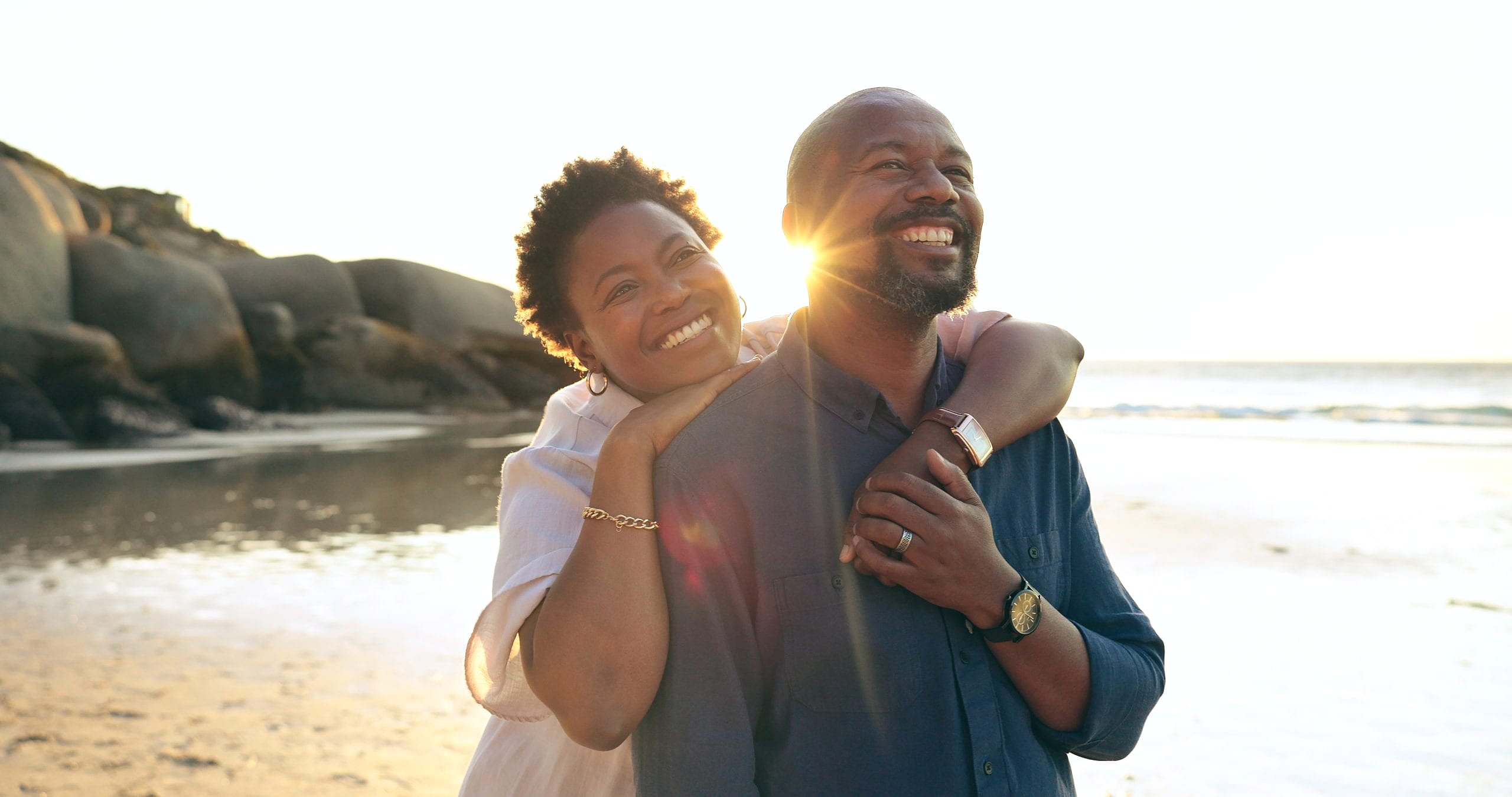 On a beach at sunset, a couple smiles as one hugs the other from behind, with rocks, the ocean, and a TMJ mouth guard case from Smile City Orthodontics in Sunnyvale, TX visible in their bag.